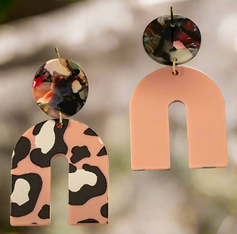 The Catalina earrings by Desiree W.A.R Store feature two abstract arch shapes: one with pink and black leopard print, the other in solid pink. Both include multicolored marble-effect round tops and gold hooks.