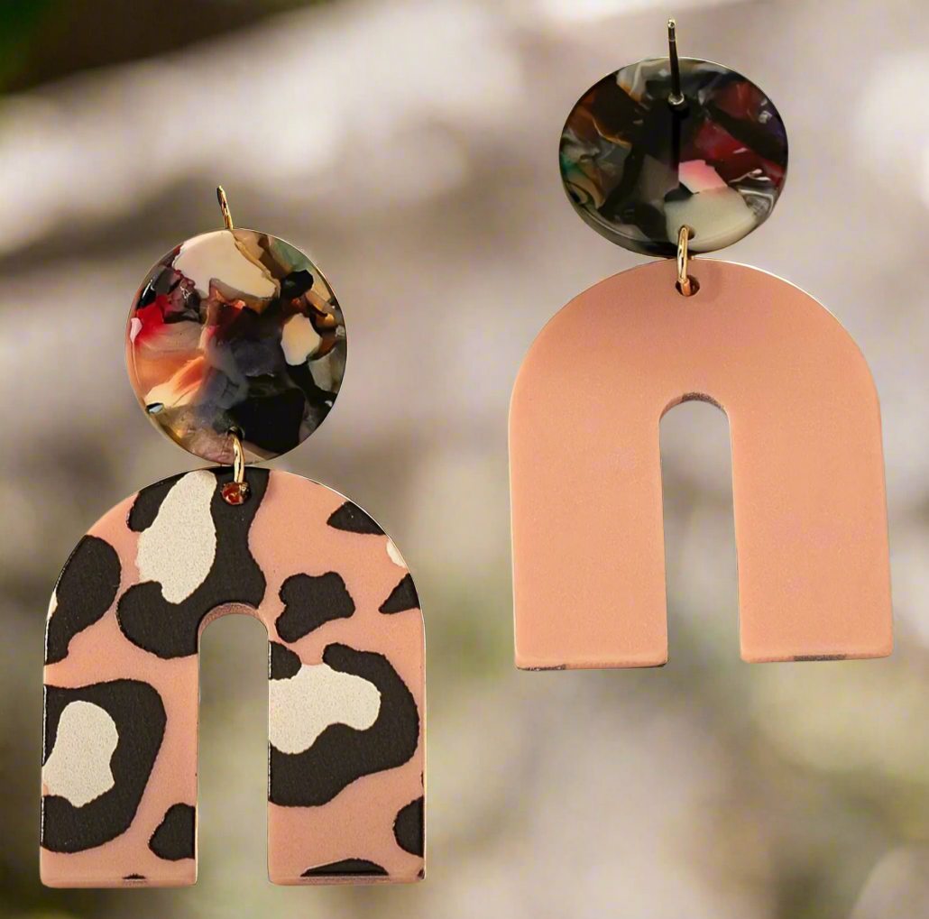 The Catalina earrings by Desiree W.A.R Store feature two abstract arch shapes: one with pink and black leopard print, the other in solid pink. Both include multicolored marble-effect round tops and gold hooks.