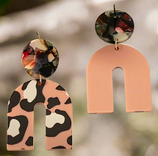 The Catalina earrings by Desiree W.A.R Store feature two abstract arch shapes: one with pink and black leopard print, the other in solid pink. Both include multicolored marble-effect round tops and gold hooks.