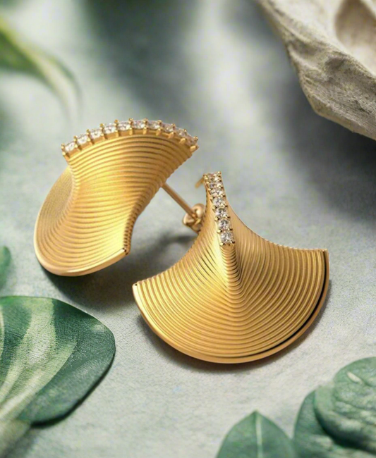 Gold fan-shaped earrings with curved ridges and a vertical row of rhinestones down the center, displayed on a neutral background with green leaf shadows.