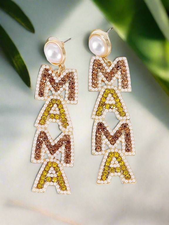 A pair of dazzling gold-plated statement earrings spelling “MAMA” in rhinestone-studded letters with pearl accents, displayed against a soft, elegant background with green leaves. Perfect for celebrating motherhood with style.