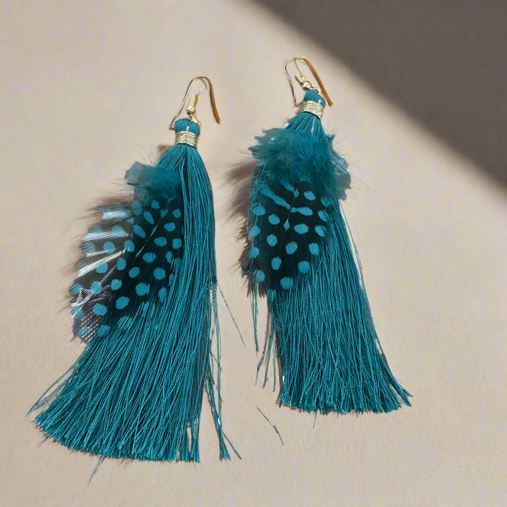 Teal feather and tassel earrings with gold thread accents, displayed on a holographic background with kitchen scene in the background.
