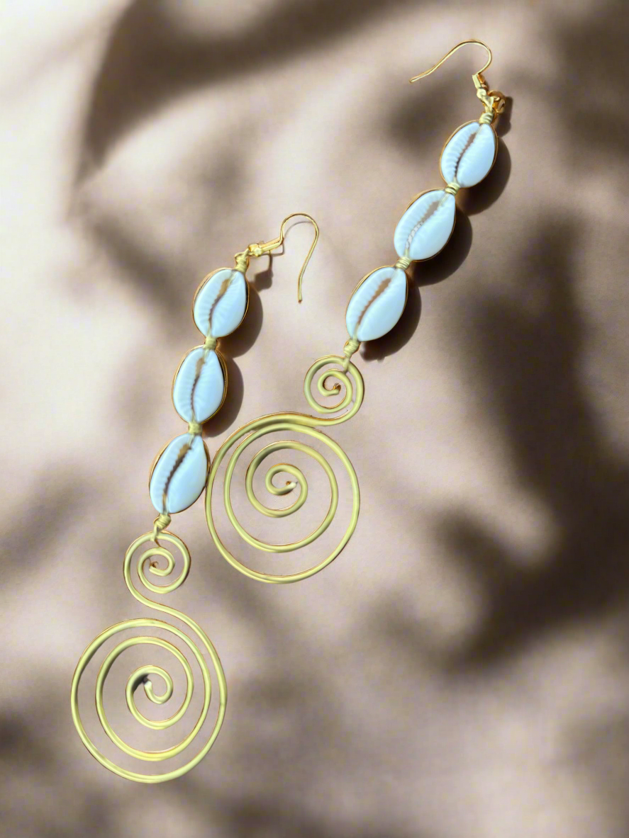 A pair of handmade dangle earrings featuring white cowrie shells connected with gold wire and finished with artistic gold spiral designs, laid out on a wooden surface.