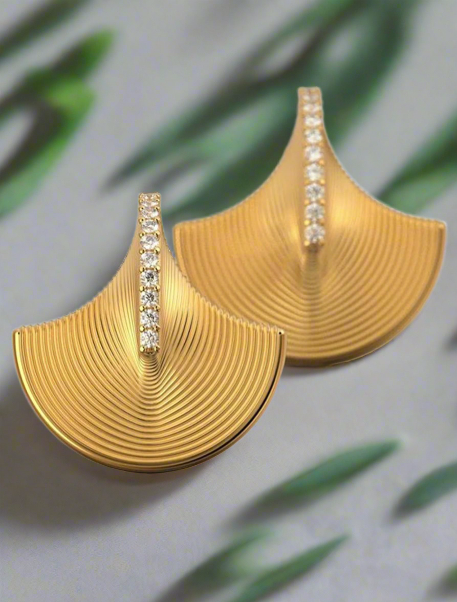 Gold fan-shaped earrings with curved ridges and a vertical row of rhinestones down the center, displayed on a neutral background with green leaf shadows.