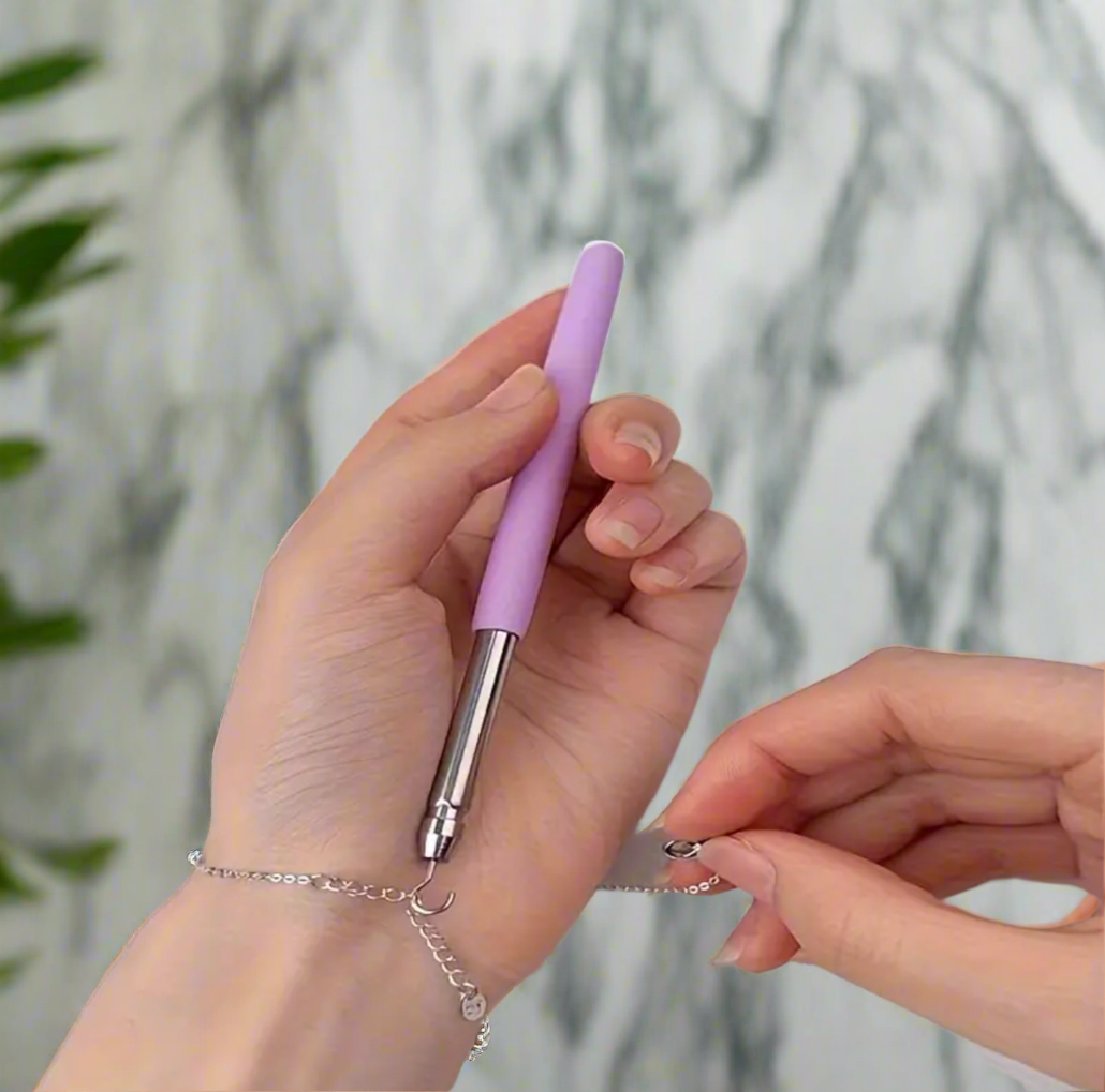 A person uses the Desiree W.A.R Stores Bracelet Fastening Hook Tool, with an ergonomic handle, to secure a clasp on their wrist. A marble-like background and green plant on the left add elegance.