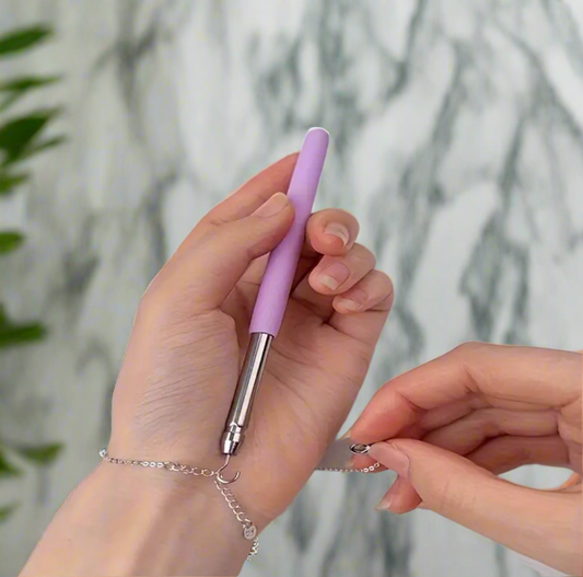 A person uses the Desiree W.A.R Stores Bracelet Fastening Hook Tool, with an ergonomic handle, to secure a clasp on their wrist. A marble-like background and green plant on the left add elegance.
