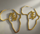 A pair of gold earrings shaped like the African continent, featuring a bold, intricate central design and smooth golden edges, displayed against a soft, neutral background.