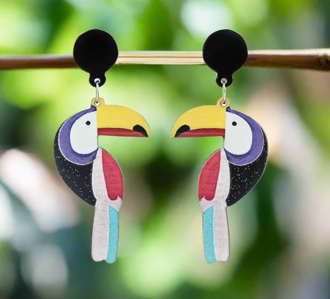 Colorful toucan-shaped statement earrings with yellow beaks, red and blue accents, and black circular tops, hanging on a display rod.
