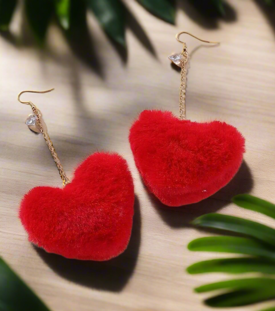 The Caprice earrings from Desiree W.A.R Store, featuring fluffy red hearts, rhinestone studs, and gold chains, rest on a wooden surface surrounded by green leaves.