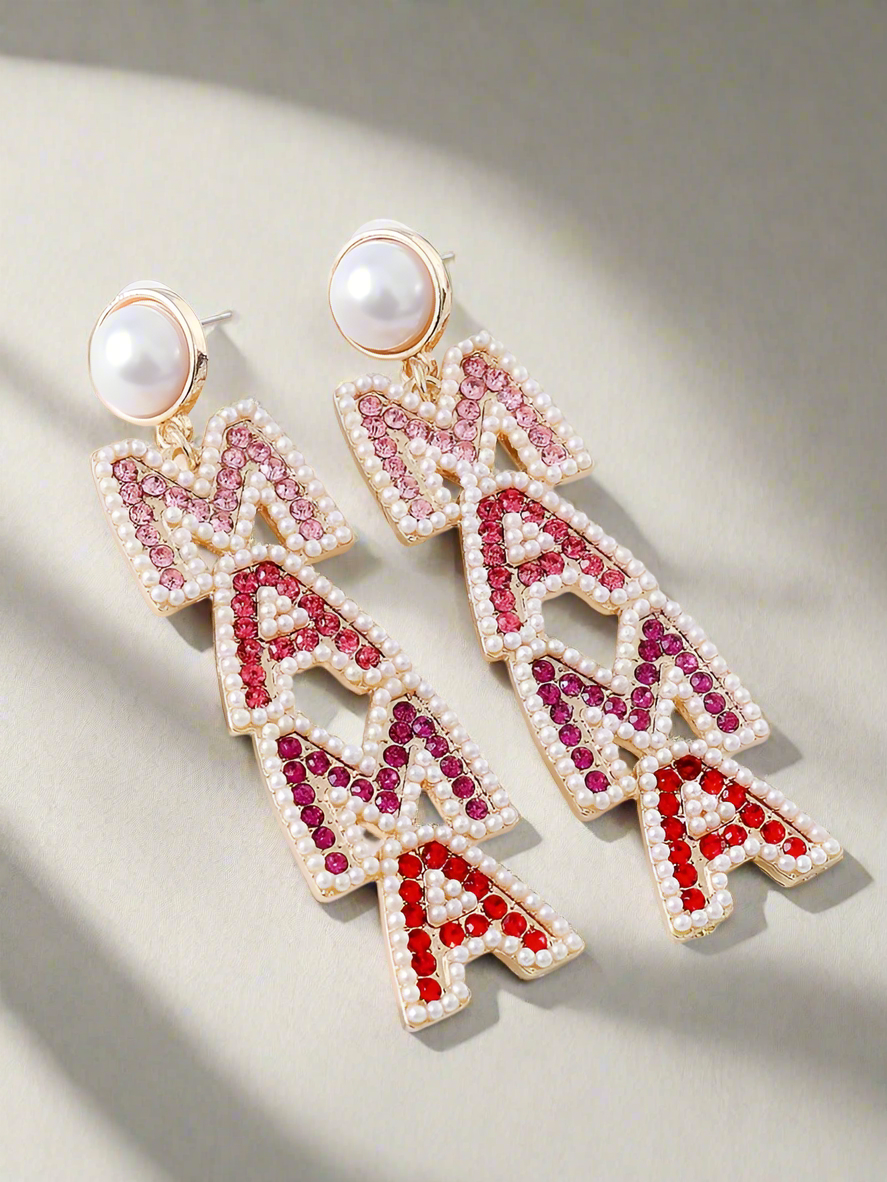 A pair of dazzling gold-plated statement earrings spelling “MAMA” in rhinestone-studded letters with pearl accents, displayed against a soft, elegant background with green leaves. Perfect for celebrating motherhood with style.