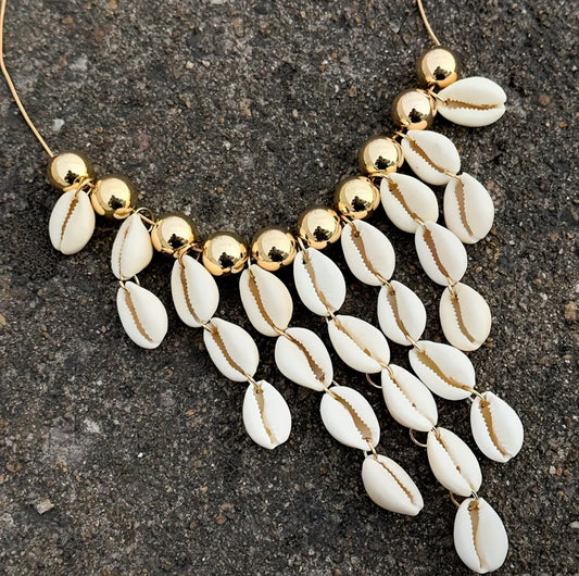 Statement necklace featuring natural white cowrie shells and shiny gold beads, displayed on a beige bust with a neutral background.