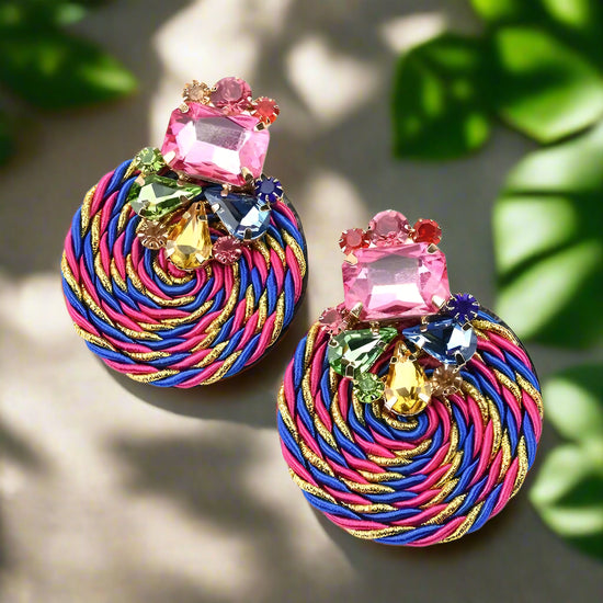 Channel vibrant island vibes with these bold, round earrings, featuring a mesmerizing swirl of braided threads and a dazzling cluster of multicolored gemstones on top. The bright pink, blue, and gold spiral creates an eye-catching centerpiece, while the sparkling crystals add a glamorous finishing touch. 