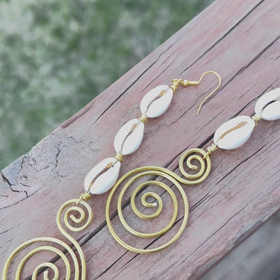 A pair of handmade dangle earrings featuring white cowrie shells connected with gold wire and finished with artistic gold spiral designs, laid out on a wooden surface.
