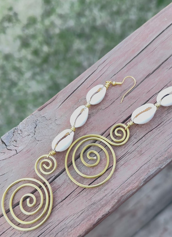 A pair of handmade dangle earrings featuring white cowrie shells connected with gold wire and finished with artistic gold spiral designs, laid out on a wooden surface.
