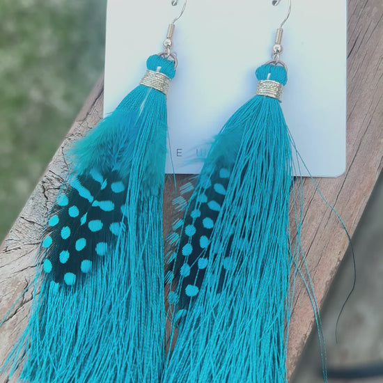 Teal feather and tassel earrings with gold thread accents, displayed on a holographic background with kitchen scene in the background.