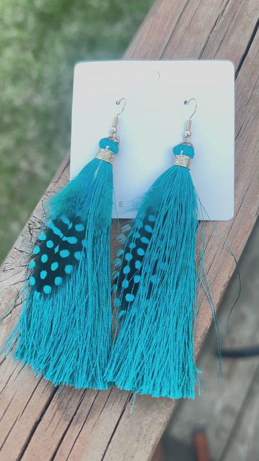 Teal feather and tassel earrings with gold thread accents, displayed on a holographic background with kitchen scene in the background.