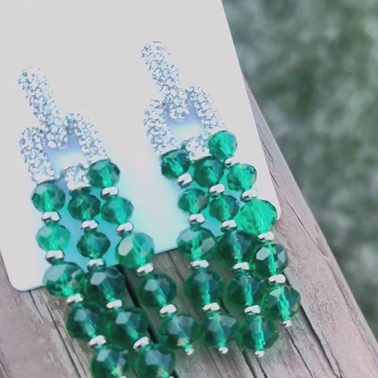 A pair of elegant drop earrings featuring cascading strands of faceted emerald green beads, accented with silver-toned spacers and topped with sparkling rhinestone-encrusted link connectors, displayed on a light surface with green palm leaves framing the image.