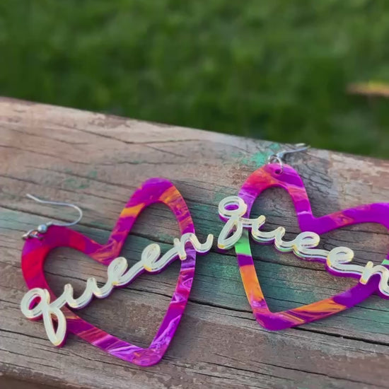 A pair of vibrant, multicolored heart-shaped earrings with the word “Queen” in gold-tone script across the center. The earrings have silver-tone hooks and feature a blend of purple, pink, orange, and green hues.